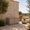 Coffre de rangement extérieur en métal taupe avec couvercle verrouillable, installé sur une terrasse en pierre aux côtés d’un salon de jardin et de plantes méditerranéennes.