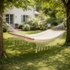 Hamac 1 place écru en coton avec franges et barres en bois suspendu entre arbres dans jardin